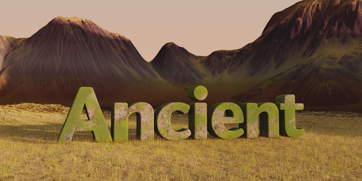 the word 'ancient' appears in a barren desert valley, surrounded by large rocky mountains. the word is partially covered in moss, obscuring its eroded rocky concrete-like texture.